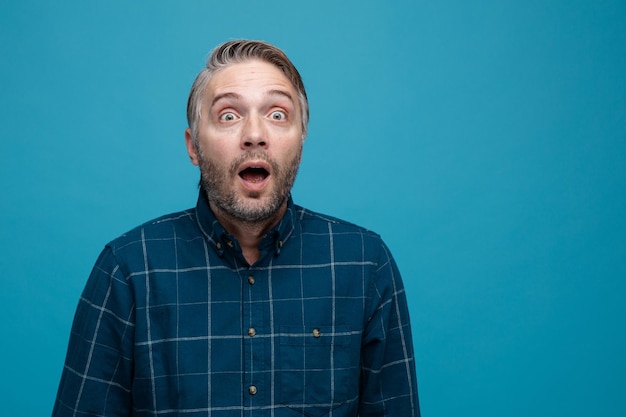 Middle age man with grey hair in dark color shirt looking aside amazed and surprised standing over blue background