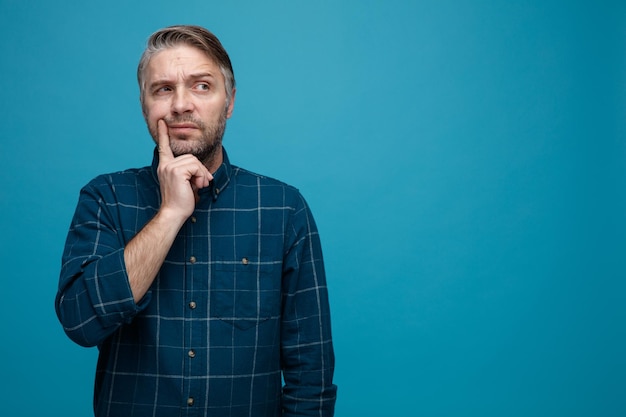 Uomo di mezza età con i capelli grigi in camicia di colore scuro che guarda da parte con un'espressione pensierosa pensando in piedi su sfondo blu