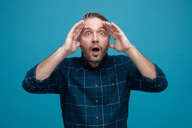 Middle age man with grey hair in dark color shirt looking at camera amazed and surprised holding hand on his head standing over blue background