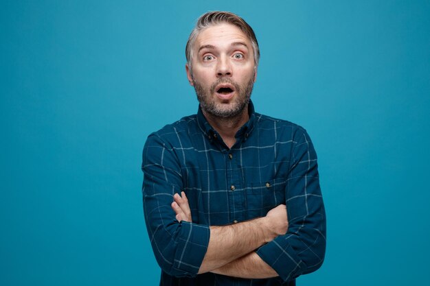 Middle age man with grey hair in dark color shirt looking at camera amazed and surprised with crossed hands on his chest standing over blue background