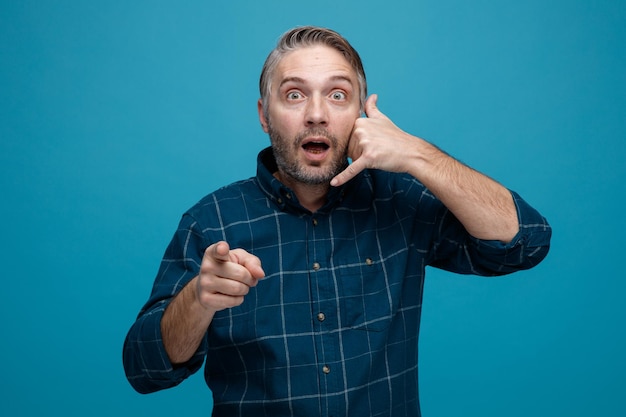 Middle age man with grey hair in dark color shirt looking at camera being surprised making call me gesture with hand pointing with index finger at camera standing over blue background