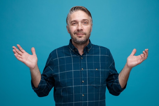 Middle age man with grey hair in dark color shirt looking at camera happy and pleased raising arms in surrender standing over blue background