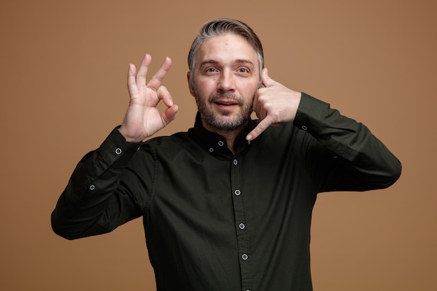 Uomo di mezza età con capelli grigi in camicia di colore scuro che guarda la fotocamera felice e positivo facendomi chiamare gesto con la mano che mostra segno ok in piedi su sfondo marrone