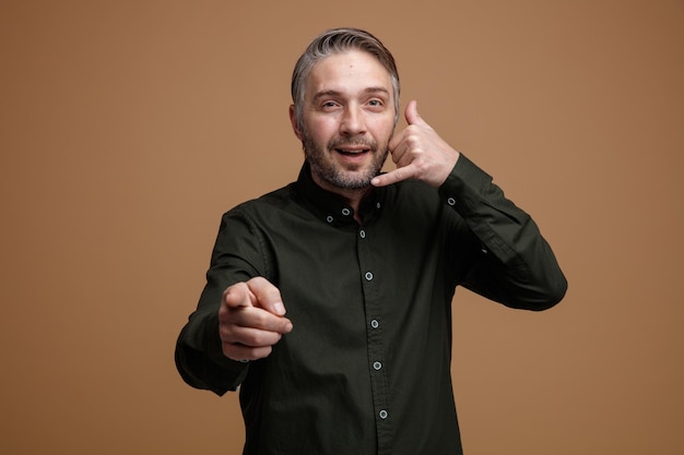 Middle age man with grey hair in dark color shirt looking at camera making call me gesture with hand pointing with index finger at camera smiling standing over brown background