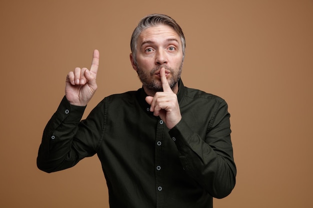 Middle age man with grey hair in dark color shirt looking at camera with serious face making silence gesture with finger on lips pointing with index finger up standing over brown background