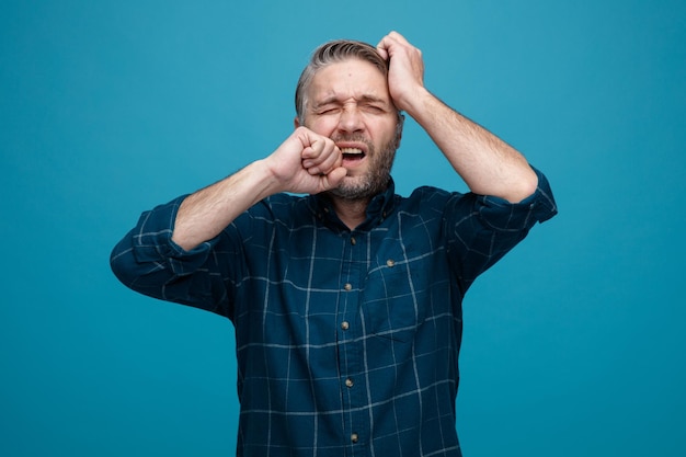 Uomo di mezza età con capelli grigi in camicia di colore scuro che sembra confuso e deluso pugno mordace in piedi su sfondo blu