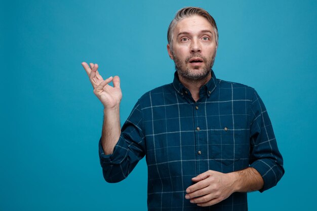 Uomo di mezza età con i capelli grigi in camicia di colore scuro che sembra confuso alzando il braccio con dubbi in piedi su sfondo blu