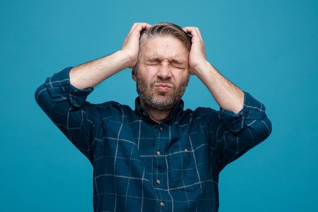 Middle age man with grey hair in dark color shirt looking unwell holding hands on his head suffering from strong headache standing over blue background