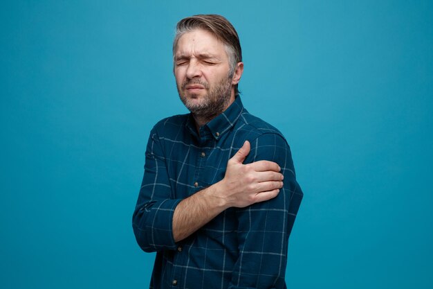 Uomo di mezza età con capelli grigi in camicia di colore scuro che sembra malato toccandosi la spalla sentendo dolore in piedi su sfondo blu