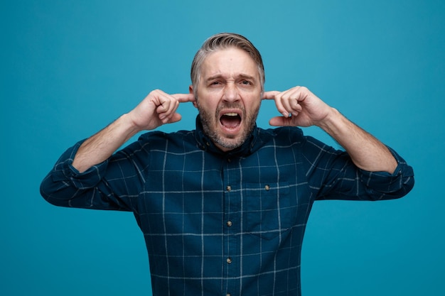 Middle age man with grey hair in dark color shirt shouting closing ears with fingers with annoyed expression standing over blue background