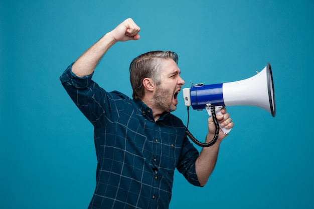 Photo middle age man with grey hair in dark color shirt shouting in megaphone being excited and angry clenching fist standing over blue background