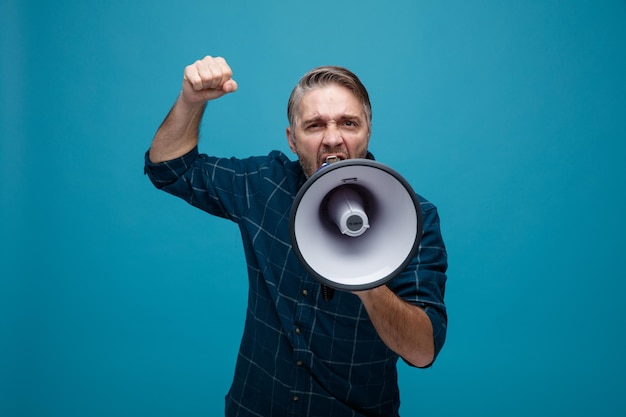 Middle age man with grey hair in dark color shirt shouting in megaphone being excited and angry raising fist standing over blue background