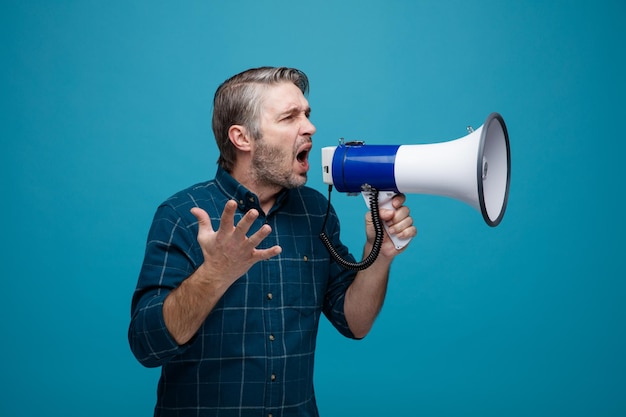 Middle age man with grey hair in dark color shirt shouting in megaphone being excited standing over blue background