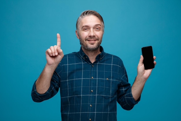 Uomo di mezza età con i capelli grigi in camicia di colore scuro che mostra lo smartphone puntato con il dito indice verso l'alto guardando la fotocamera sorridente fiducioso in piedi su sfondo blu