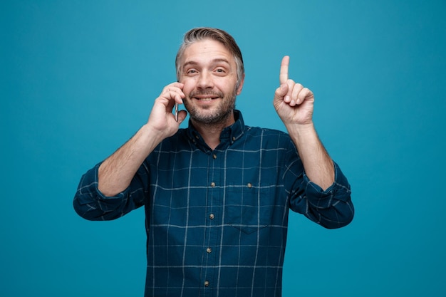 Middle age man with grey hair in dark color shirt talking on mobile phone looking at camera smiling cheerfully showing index finger having new idea standing over blue background