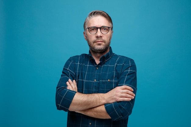 Middle age man with grey hair in dark color shirt wearing glasses looking at camera frowningly standing over blue background