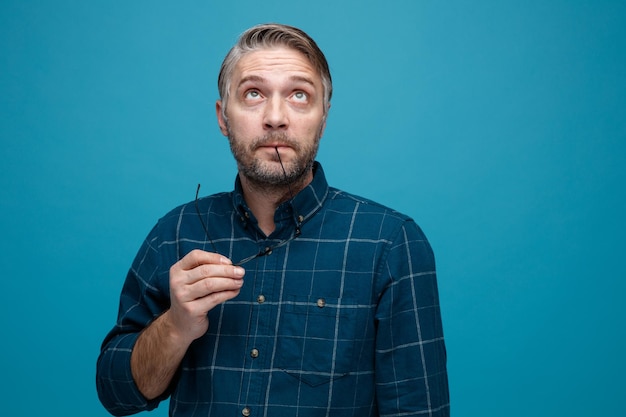 Middle age man with grey hair in dark color shirt wearing glasses looking up puzzled standing over blue background