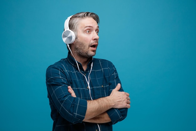 Middle age man with grey hair in dark color shirt with headphones looking aside being surprised with arms crossed on his chest standing over blue background