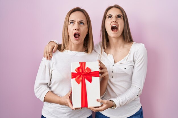 Middle age mother and young daughter holding mothers day gift angry and mad screaming frustrated and furious shouting with anger looking up