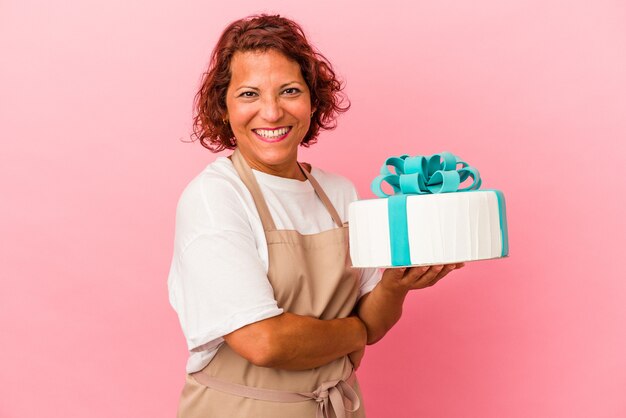 Middle age pastry latin woman holding a cake isolated on pink background laughing and having fun.