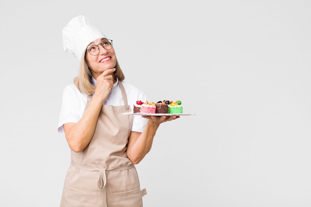 Middle age pretty baker  woman with cakes against copy space wall