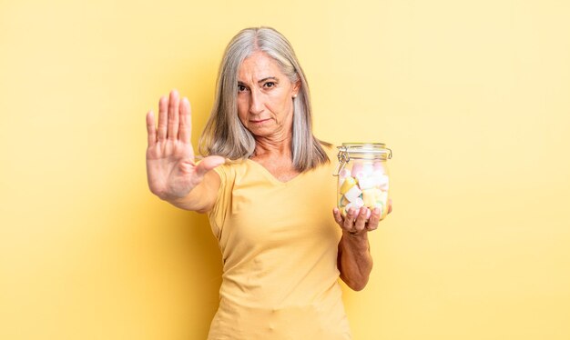 Middle age pretty woman looking serious showing open palm making stop gesture. candies bottle concept