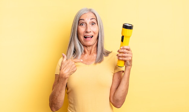 Middle age pretty woman looking shocked and surprised with mouth wide open, pointing to self. flashlight concept