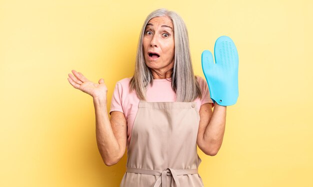 Middle age pretty woman looking surprised and shocked, with jaw dropped holding an object. oven mitt concept