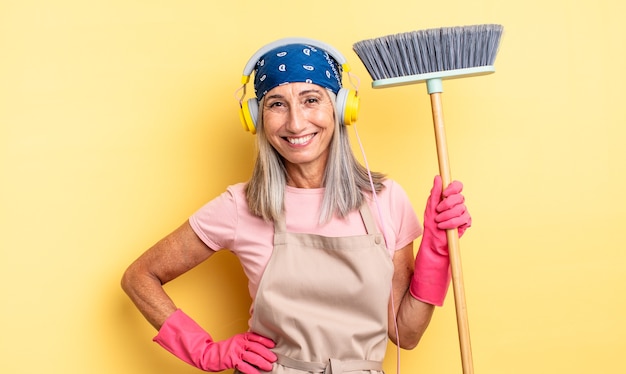 Middle age pretty woman smiling happily with a hand on hip and confident. household and broom concept