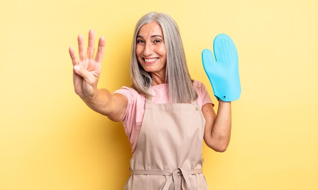 Middle age pretty woman smiling and looking friendly, showing number four. oven mitt concept