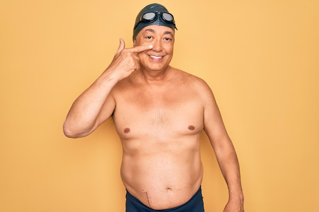 Middle age senior greyhaired swimmer man wearing swimsuit cap and goggles Pointing with hand finger to face and nose smiling cheerful Beauty concept