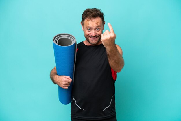 Middle age sport man going to yoga classes while holding a mat isolated on blue background doing coming gesture