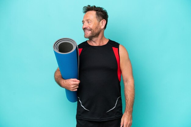 Middle age sport man going to yoga classes while holding a mat isolated on blue background looking side