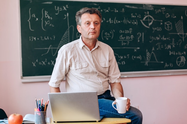 Middle age teacher holding a cup sitting next an open laptop  on the table.