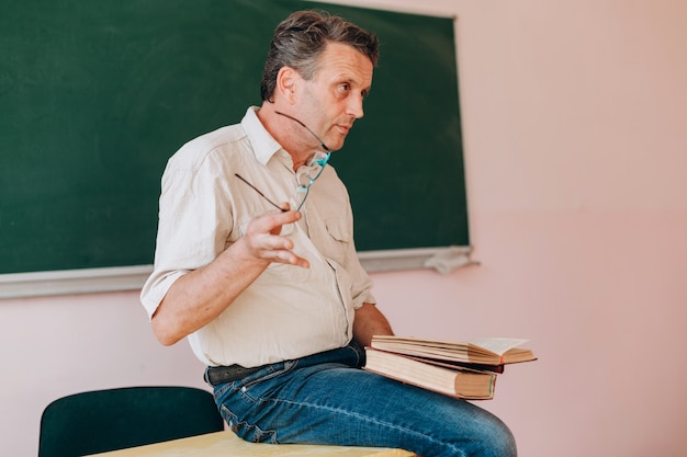 Middle age teacher holding a glasses and sitting with open textbook.