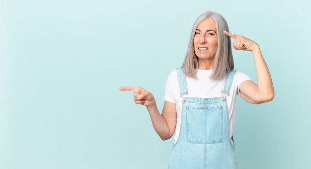 Donna di mezza età con i capelli bianchi che si sente confusa e perplessa, mostra che sei pazzo e indica il lato