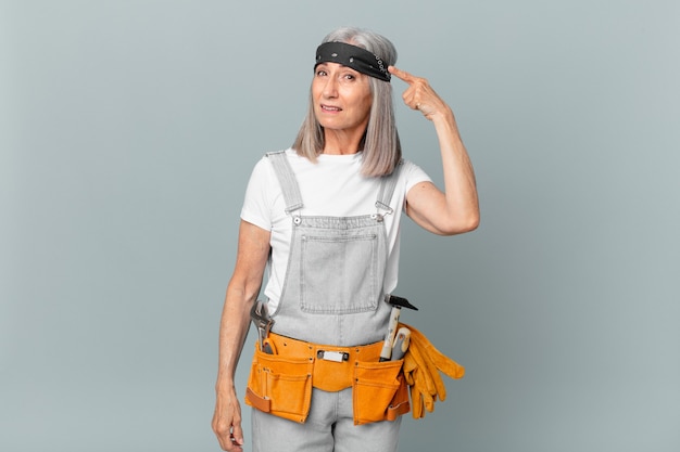 Middle age white hair woman feeling confused and puzzled, showing you are insane and wearing work wear and tools. housekeeping concept