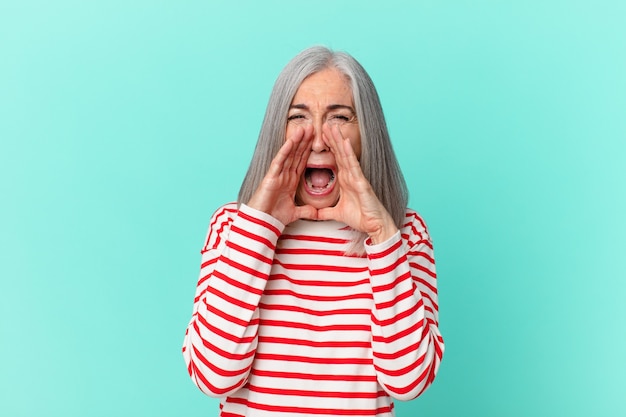 Middle age white hair woman feeling happy,giving a big shout out with hands next to mouth