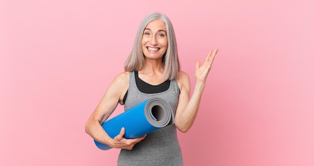 Donna di mezza età con i capelli bianchi che si sente felice, sorpresa di realizzare una soluzione o un'idea e con in mano un tappetino da yoga. concetto di fitness