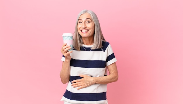 Женщина средних лет с белыми волосами громко смеется над какой-то веселой шуткой и держит контейнер для кофе на вынос