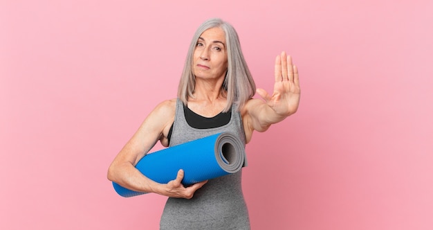 Женщина средних лет с белыми волосами выглядит серьезной, показывая открытую ладонь, делая стоп-жест и держа коврик для йоги. фитнес-концепция