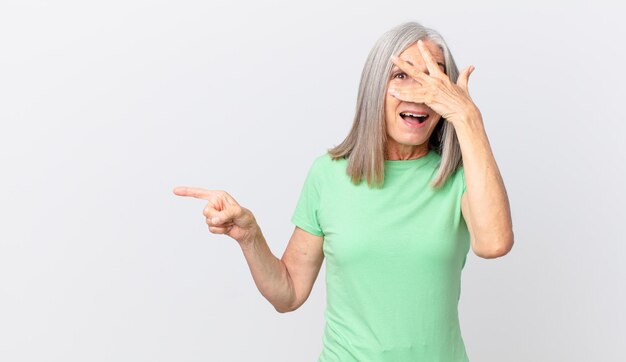 Middle age white hair woman looking shocked, scared or terrified, covering face with hand and pointing to the side