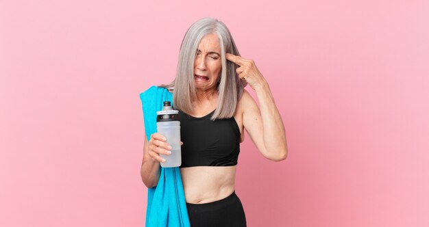 Middle age white hair woman looking unhappy and stressed, suicide gesture making gun sign with a towel and water bottle. fitness concept