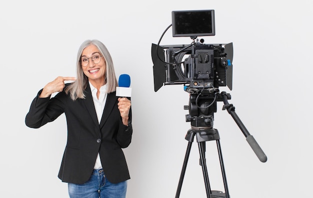 Middle age white hair woman smiling confidently pointing to own broad smile and holding a microphone. television presenter concept