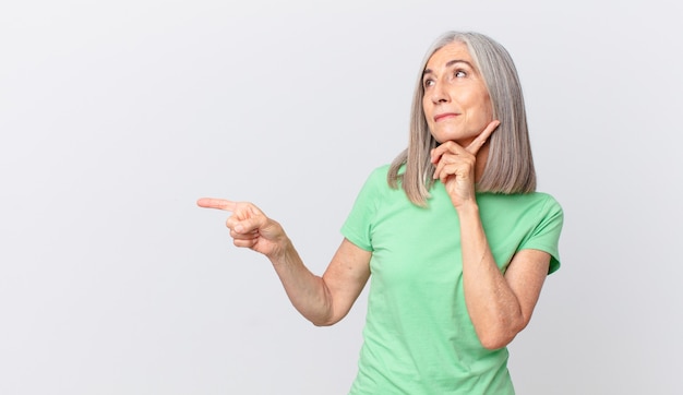 Middle age white hair woman smiling happily and daydreaming or doubting and pointing to the side