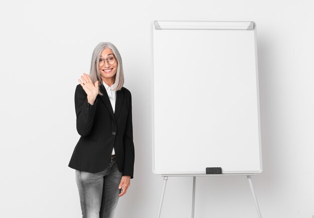 Middle age white hair woman smiling happily, waving hand, welcoming and greeting you and a board copy space. business concept