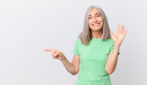 Middle age white hair woman smiling happily, waving hand, welcoming and greeting you and pointing to the side