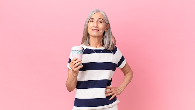 Donna di mezza età con i capelli bianchi che sorride felicemente con una mano sull'anca e sicura e tiene in mano un contenitore di caffè da asporto