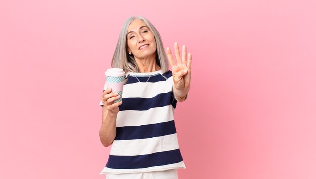 Donna di mezza età con i capelli bianchi che sorride e sembra amichevole, mostra il numero quattro e tiene in mano un contenitore di caffè da asporto