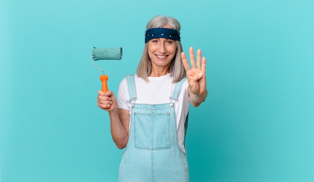 Middle age white hair woman smiling and looking friendly, showing number four with a roller painting a wall